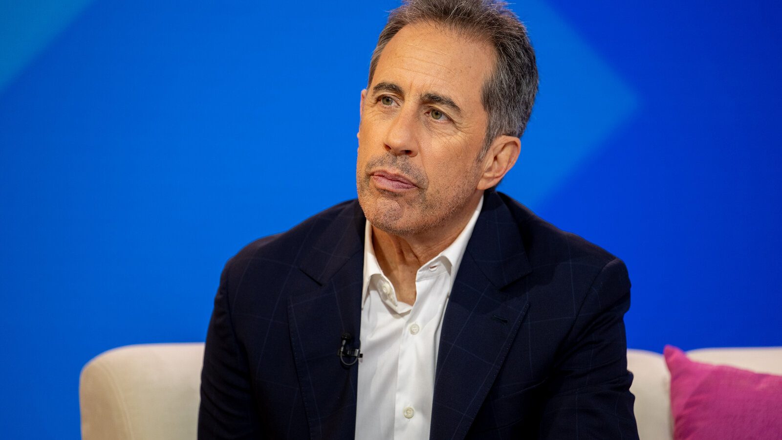 Jerry Seinfeld in a suit is seated on a couch, looking thoughtfully to the side