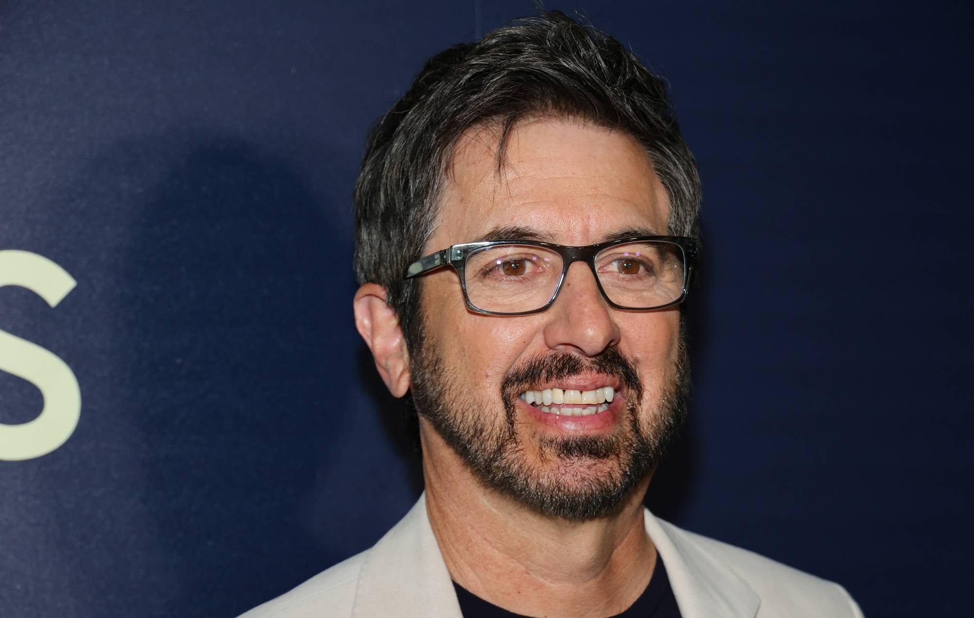 Ray Romano wearing glasses and a white jacket stands confidently