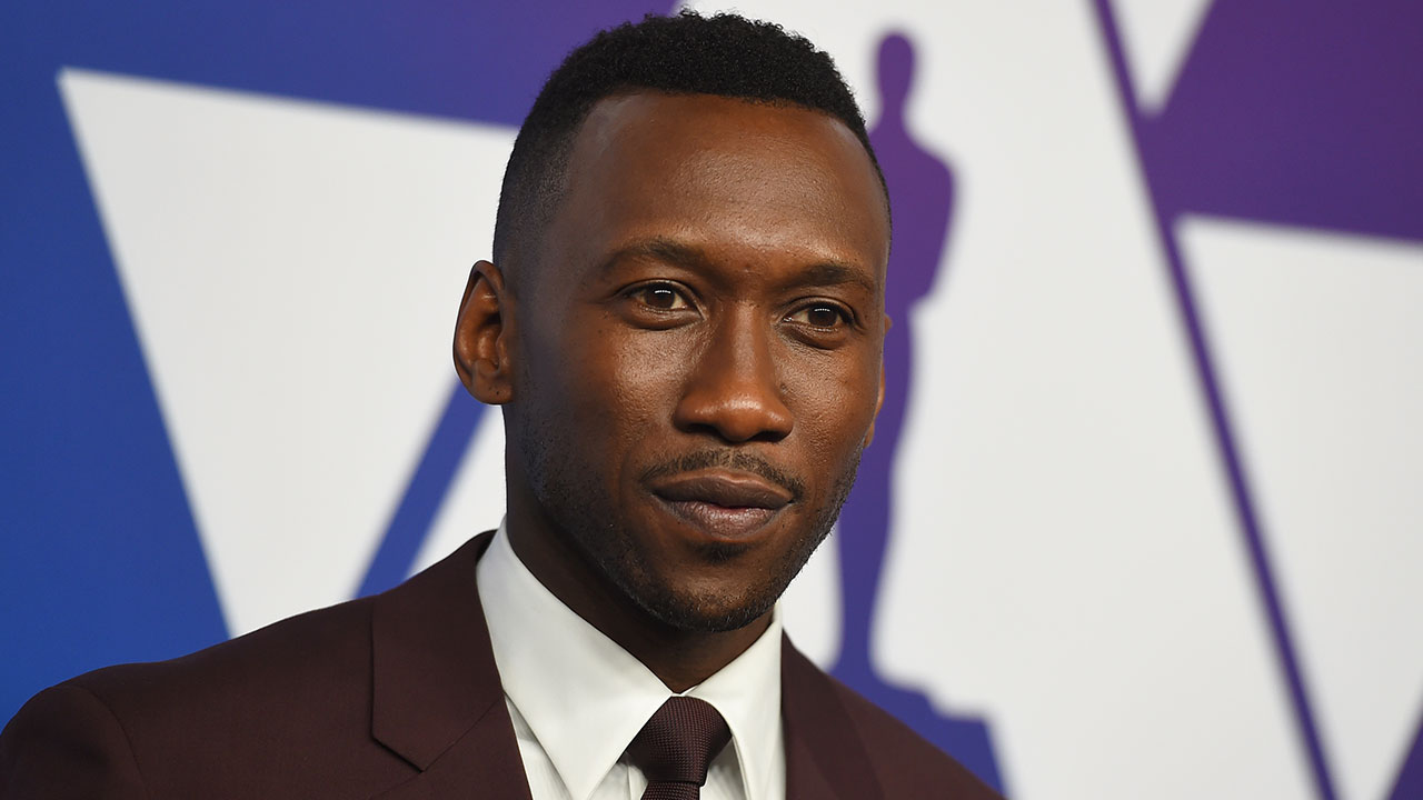 Mahershala Ali in a suit and tie stands against a blue and white background